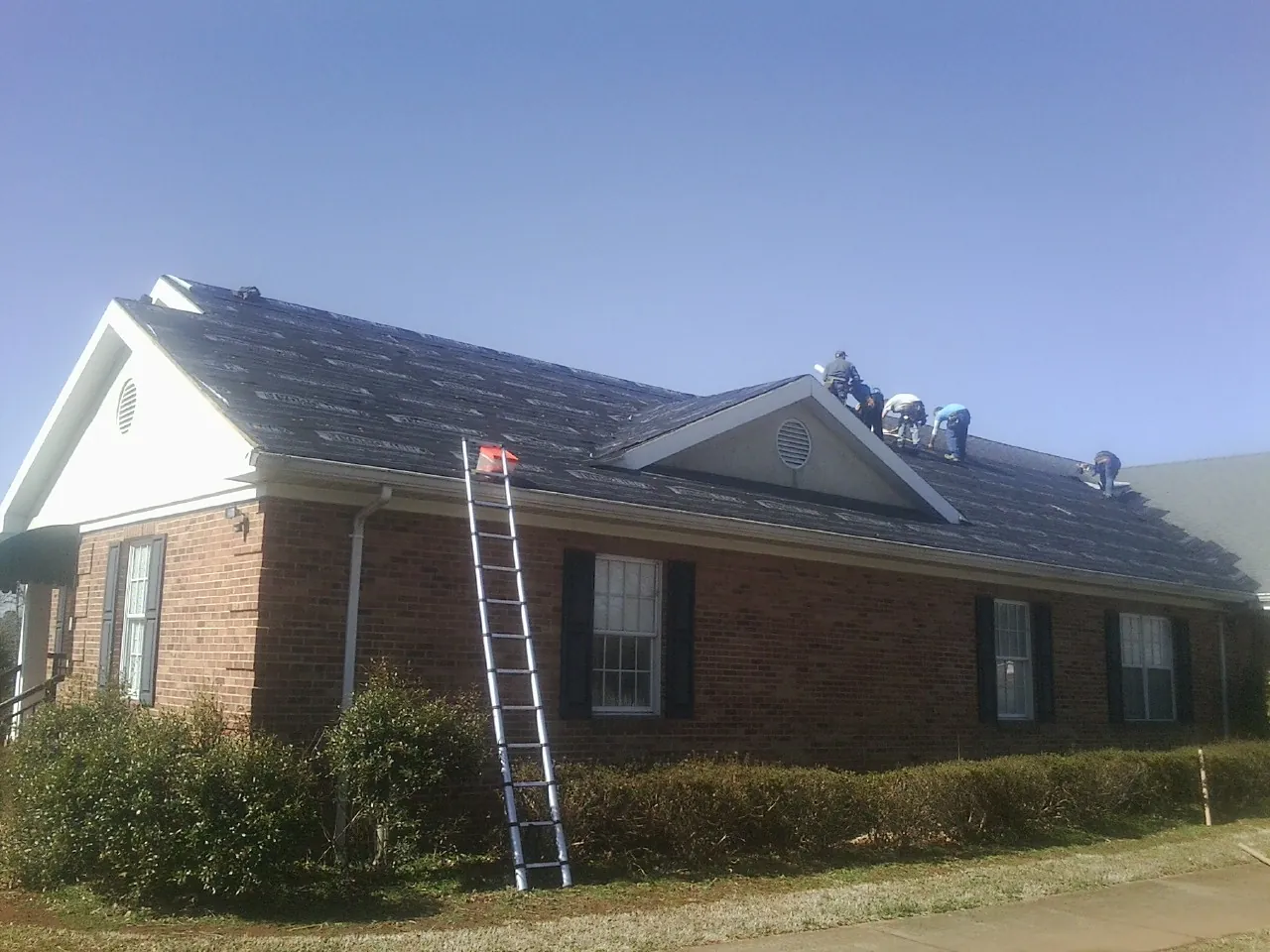 Crew tearing shingles off a brick ranch roof with a ladder set — Emergency Roof Repair work by our crew in War