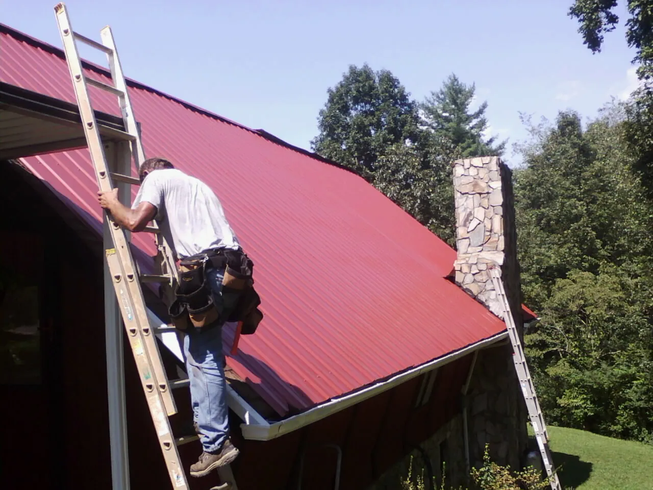 Roofer on a ladder beside a red metal roof and stone chimney — Soffit & Fascia Repair by Roofing Brothers in War